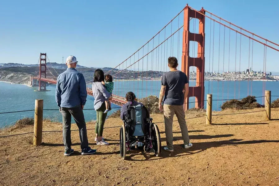 Um grupo de pessoas, incluindo uma pessoa em uma cadeira de rodas, é visto de trás enquanto olham para a Golden Gate Bridge de Marin Headlands.